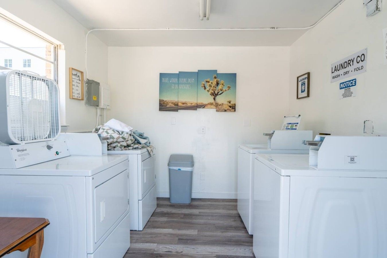 Een wasserette met twee Whirlpool wasmachines, een droger, een prullenbak en een ventilator. Er hangen drie ingelijste foto's aan de muur en een bord met de naam van een wasserij.