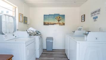 Een wasserette met twee Whirlpool wasmachines, een droger, een prullenbak en een ventilator. Er hangen drie ingelijste foto's aan de muur en een bord met de naam van een wasserij.