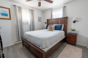 Une chambre à coucher avec un lit en bois, une table de chevet, un ventilateur de plafond et une fenêtre avec des rideaux.