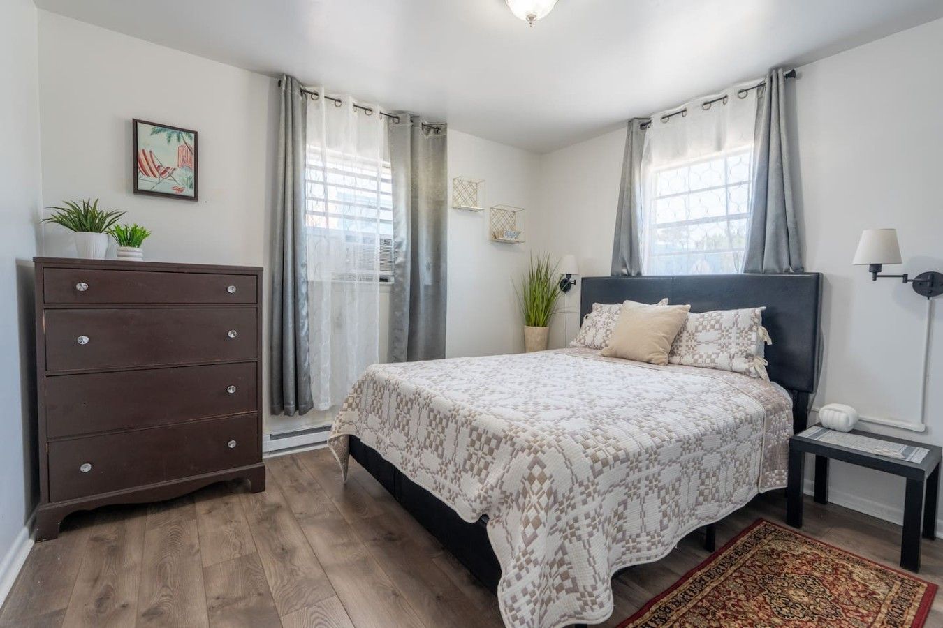 Een slaapkamer met een bed, een dressoir, een nachtkastje, een lamp en een raam met gordijnen.
