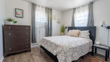 Een slaapkamer met een bed, een dressoir, een nachtkastje, een lamp en een raam met gordijnen.