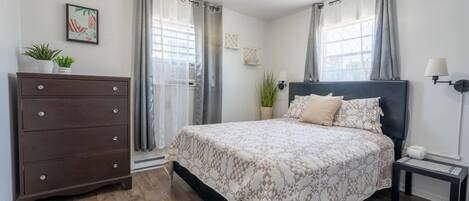 A bedroom with a bed, dresser, nightstand, lamp, and a window with curtains.