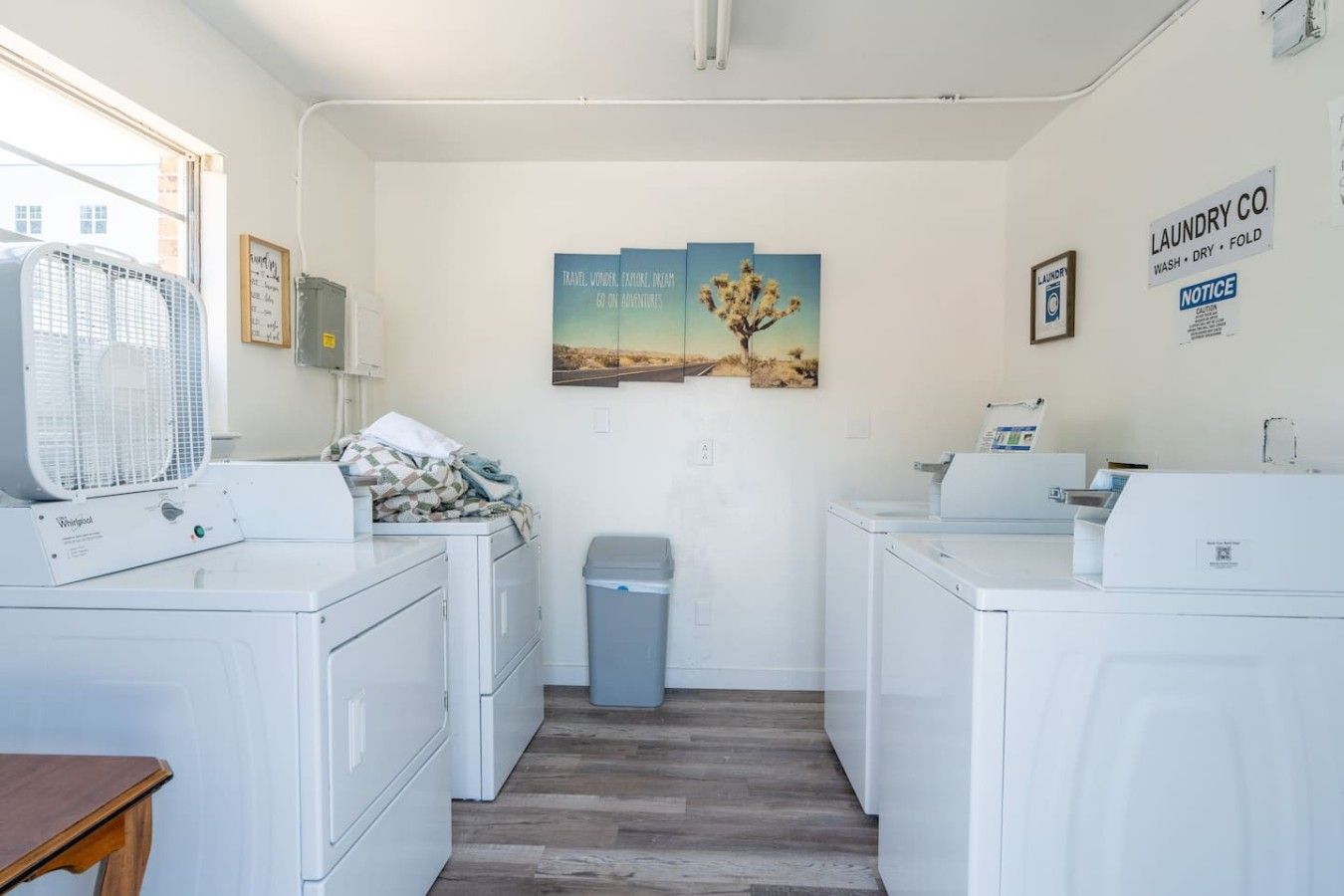 A laundry room with two Whirlpool washers, a dryer, a trash bin, and a fan. There are three framed pictures on the wall and a sign for the laundry co.
