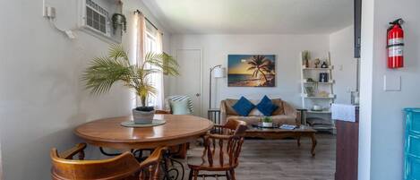 A dining area with a round wooden table and chairs, a potted plant, and a painting of a beach scene.