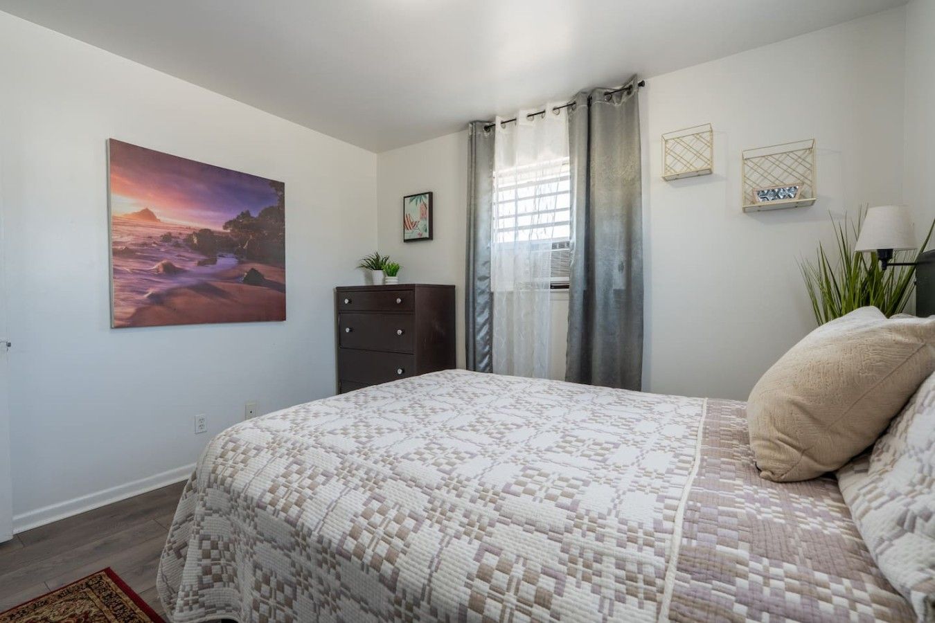 Un dormitorio con cama, cómoda, ventana con cortinas, un cuadro y una planta.