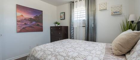 Un dormitorio con cama, cómoda, ventana con cortinas, un cuadro y una planta.