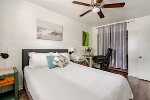 A hotel room with a bed, a desk, a chair, a ceiling fan, a painting, and a curtain. - Mystique Blue 2 BR and Bath APT near Beach (Virginia Beach)