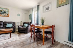 A living room with a sofa, a coffee table, a dining table with chairs, a potted plant, and a wall-mounted air conditioning unit. - Mystique Blue 2 BR and Bath APT near Beach (Virginia Beach)