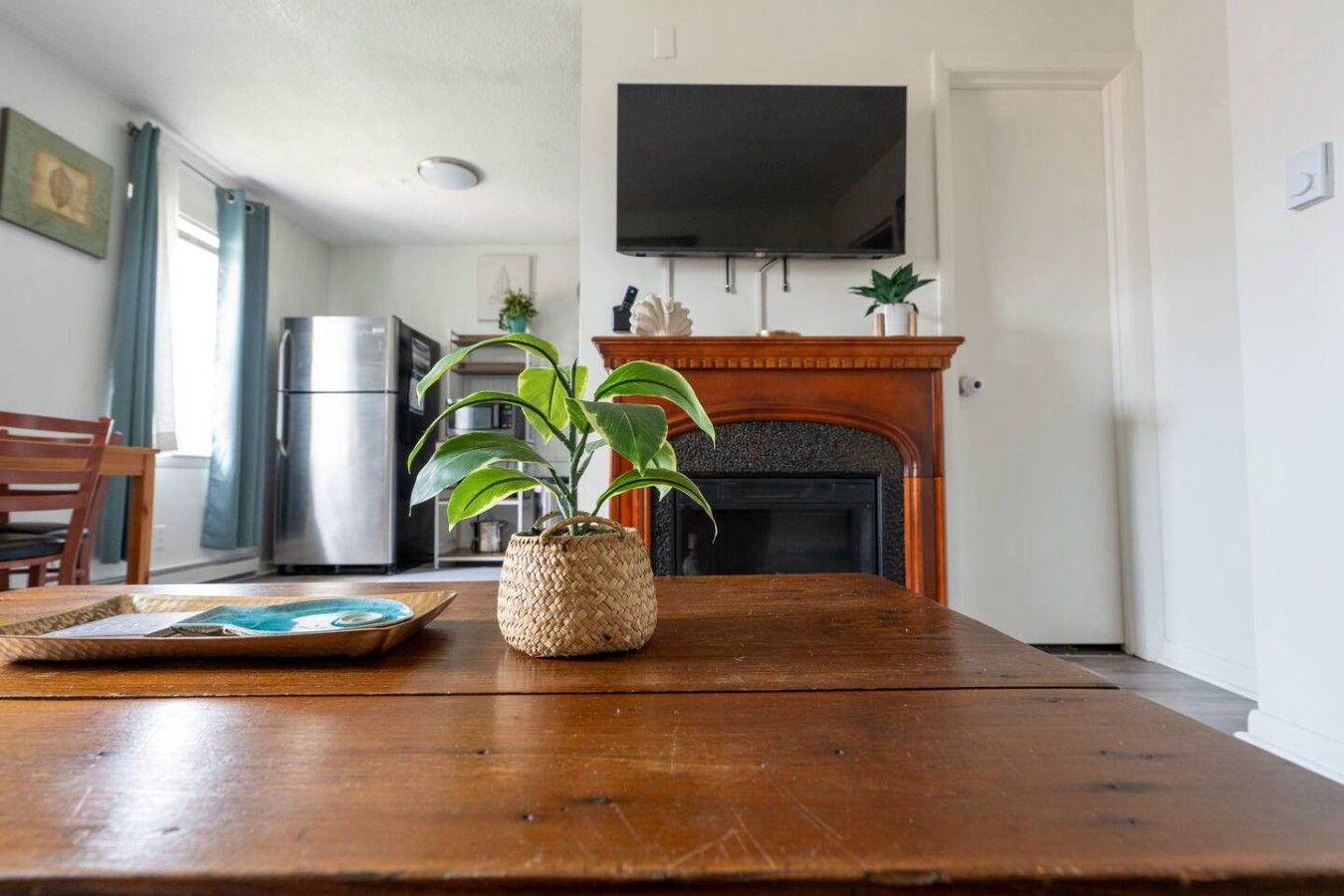 A wooden dining table with a woven basket and a placemat, a television mounted above a fireplace, and a kitchen area with a refrigerator and cabinets in the background.