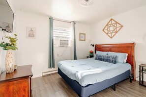 A bedroom with a bed, a wooden headboard, a nightstand, a wall-mounted artwork, and a window with curtains. - Oceanfront 2BR • Walk to Atlantic Park (Virginia Beach)