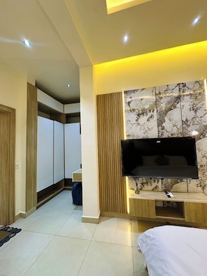 Una habitación de hotel moderna con una televisión de pantalla plana montada en un panel de madera, un espejo grande y una cama con cubrecama blanco.
