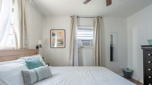 Ein Hotelzimmer mit einem Bett, einem Nachttisch, einer Lampe, einem Fenster mit Vorhängen, einer Klimaanlage, einem Schminktisch und einem an der Wand befestigten Spiegel.