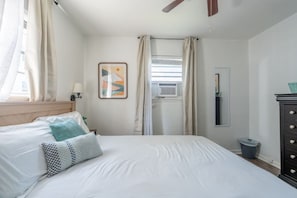 A hotel room with a bed, a nightstand, a lamp, a window with curtains, an air conditioning unit, a dresser, and a wall-mounted mirror. - Quiet 2BR • Walk to Beach, ViBe & The Dome (Virginia Beach)