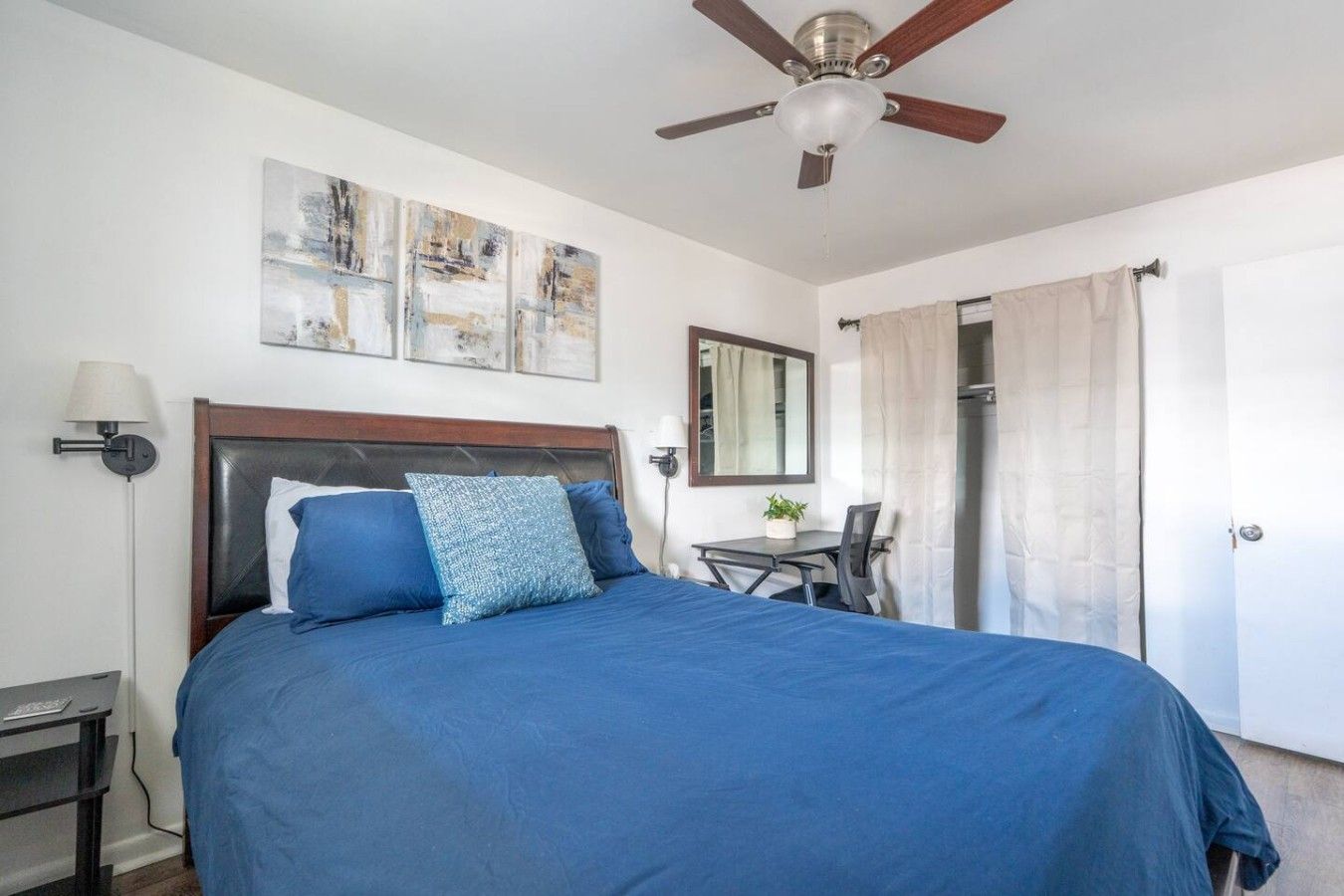 Une chambre à coucher comprenant un lit, un bureau, une chaise, un ventilateur de plafond et des œuvres d’art accrochées au mur.