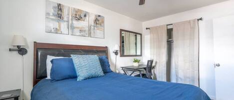 Une chambre à coucher comprenant un lit, un bureau, une chaise, un ventilateur de plafond et des œuvres d’art accrochées au mur.