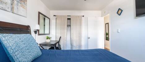 Quarto com cama, escrivaninha, cadeira, ventoinha no teto e televisão na parede.