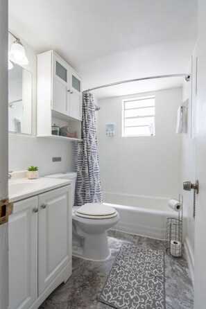 A bathroom with a toilet, a bathtub, a shower curtain, a window, and a sink with a mirror above it. - Quiet 2BR • Walk to Beach, ViBe & The Dome (Virginia Beach)
