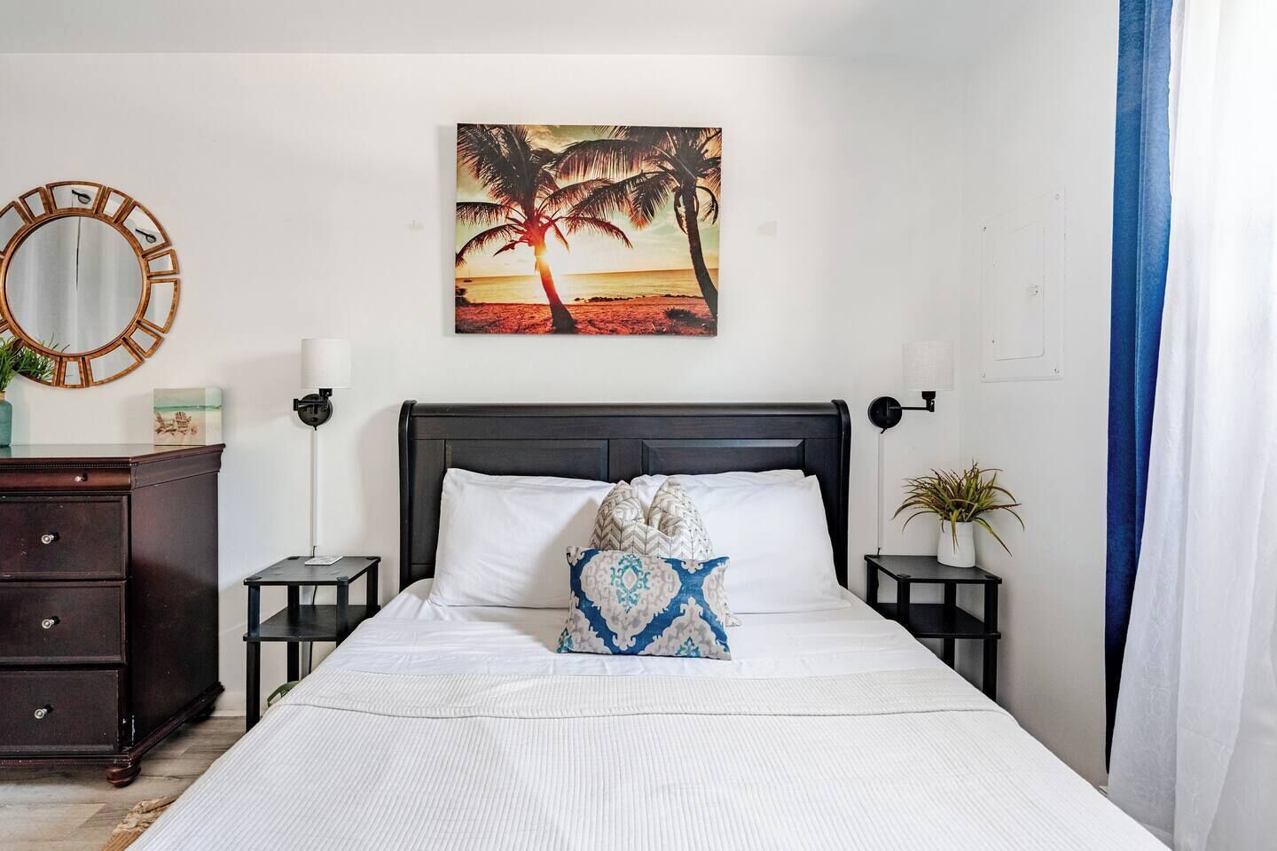 A hotel room with a bed, a nightstand, a dresser, a wall-mounted mirror, and a painting of a sunset.