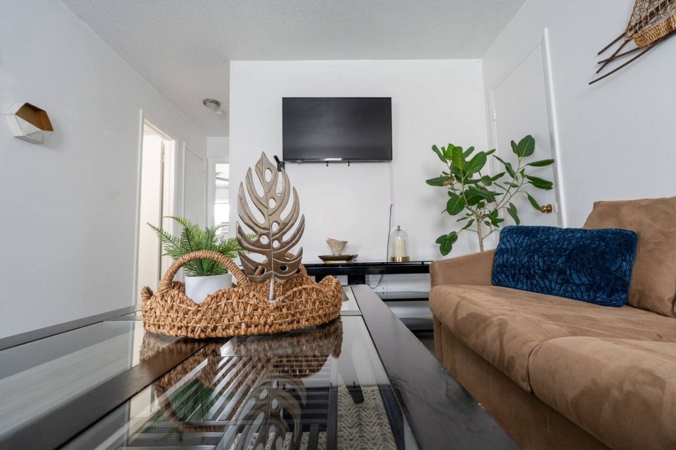 Een moderne woonkamer met een glazen salontafel, een bruine bank, een blauw kussen, een aan de muur bevestigde televisie en een potplant.