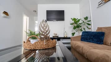Een moderne woonkamer met een glazen salontafel, een bruine bank, een blauw kussen, een aan de muur bevestigde televisie en een potplant.