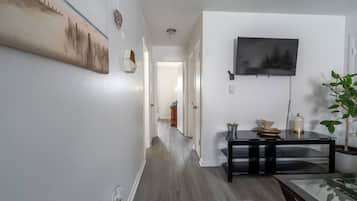 Um corredor com piso de madeira, uma mesa preta com objetos em cima, uma televisão montada na parede e um quadro na parede.