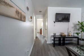 Un pasillo con piso de madera, una mesa negra con objetos encima, un televisor montado y un cuadro en la pared.