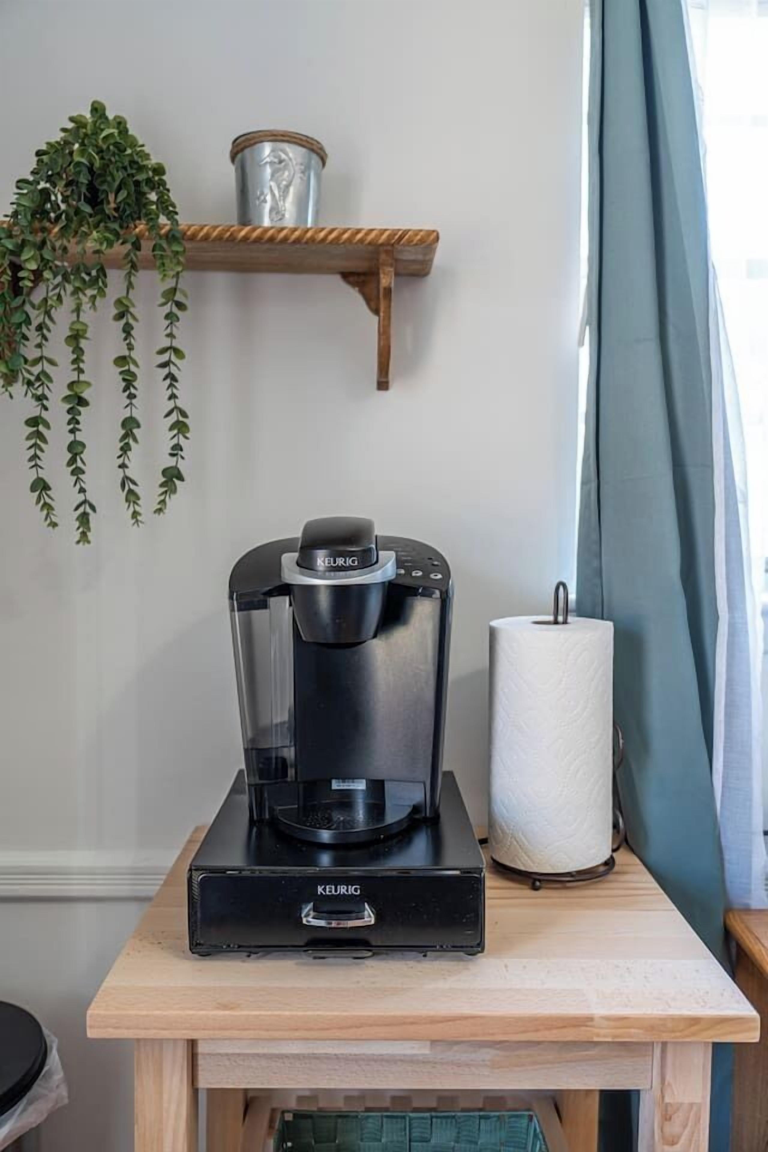 A Keurig coffee maker on a wooden table with a hanging plant and a metal container on a shelf.