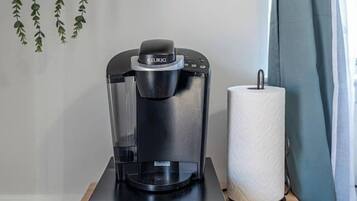A Keurig coffee maker on a wooden table with a hanging plant and a metal container on a shelf.