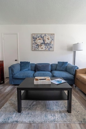 Ein Wohnzimmer mit einem dunkelholzigen Couchtisch, einem blauen Sofa und einem beigefarbenen Sessel. Über dem Sofa hängt ein Gemälde.