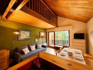 Ein gemütliches Zimmer mit Holzbalkendecke, einer grünen Wand, einem Sofa mit Kissen, einem Esstisch für zwei Personen und einem an der Wand befestigten Fernseher.