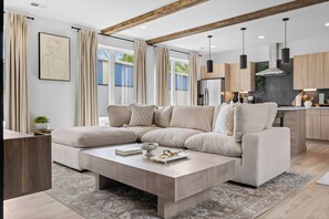 Ein modernes Wohnzimmer mit einem Ecksofa, einem Couchtisch und einer Küchenzeile mit Holzschränken und Pendelleuchten.