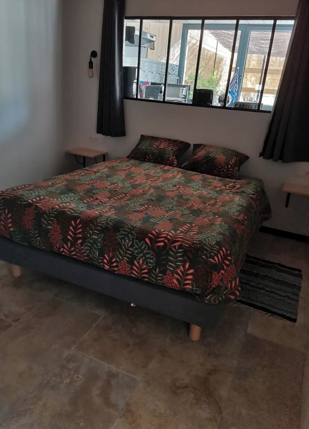 Un dormitorio con cama, dos mesitas de noche, ventana con vistas y cortina.