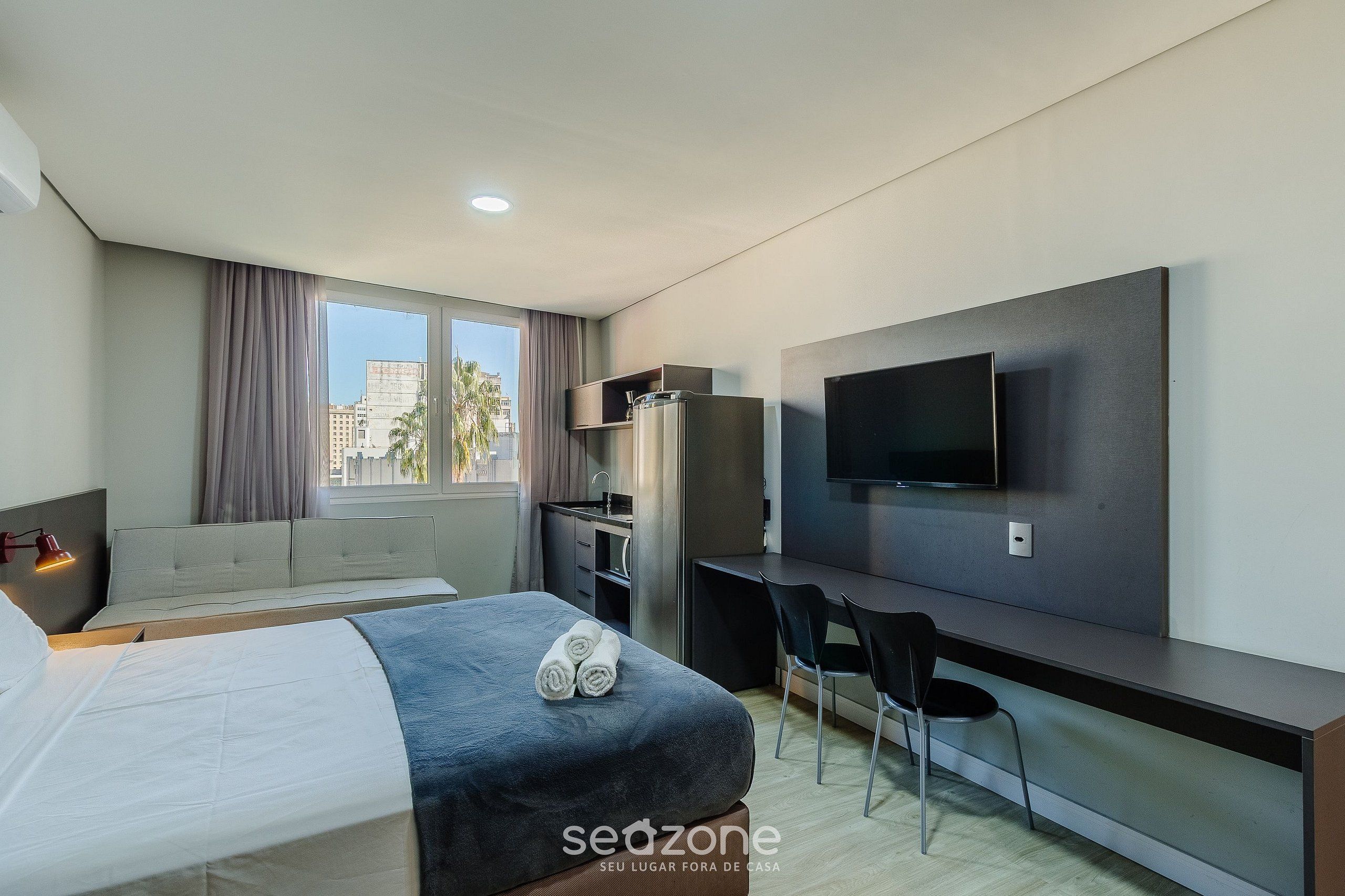 A modern hotel room with a bed, a desk, a chair, a TV, and a window with curtains.