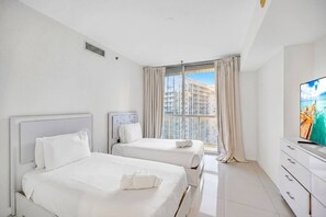Ein Hotelzimmer mit zwei Betten, einem großen Fenster mit Vorhängen, einem Flachbildfernseher und einer weißen Kommode.