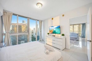 Ein modernes Hotelzimmer mit einem großen Bett, einem Flachbildfernseher, Blick auf die Stadt und einem Balkon mit Vorhängen.