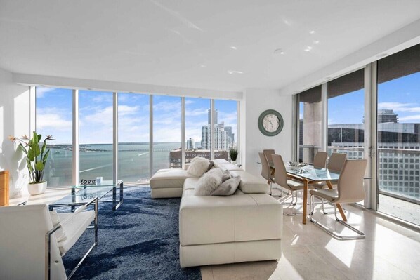 Ein modernes Wohnzimmer mit großem Fenster, einer Couch, einem Esstisch mit Stühlen und Blick auf Stadt und Meer.