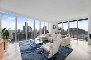 Ein modernes Wohnzimmer mit einer großen Couch, einem Glastisch und Blick auf die Skyline der Stadt durch raumhohe Fenster.
