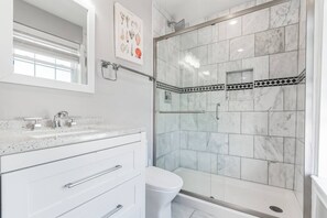 Een badkamer met een glazen douchecabine, witte marmeren tegels, een wit wastafelmeubel met een puntpatroon en een ingelijste foto aan de muur.