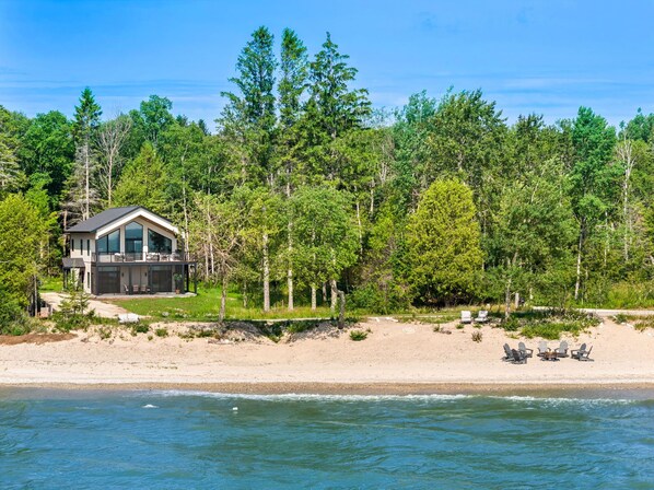 A modern beach house with a large deck, surrounded by dense forest, and a sandy beach with beach chairs.