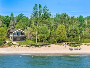 Una moderna casa de playa con una amplia terraza, rodeada de un bosque frondoso y una playa de arena con sillas.