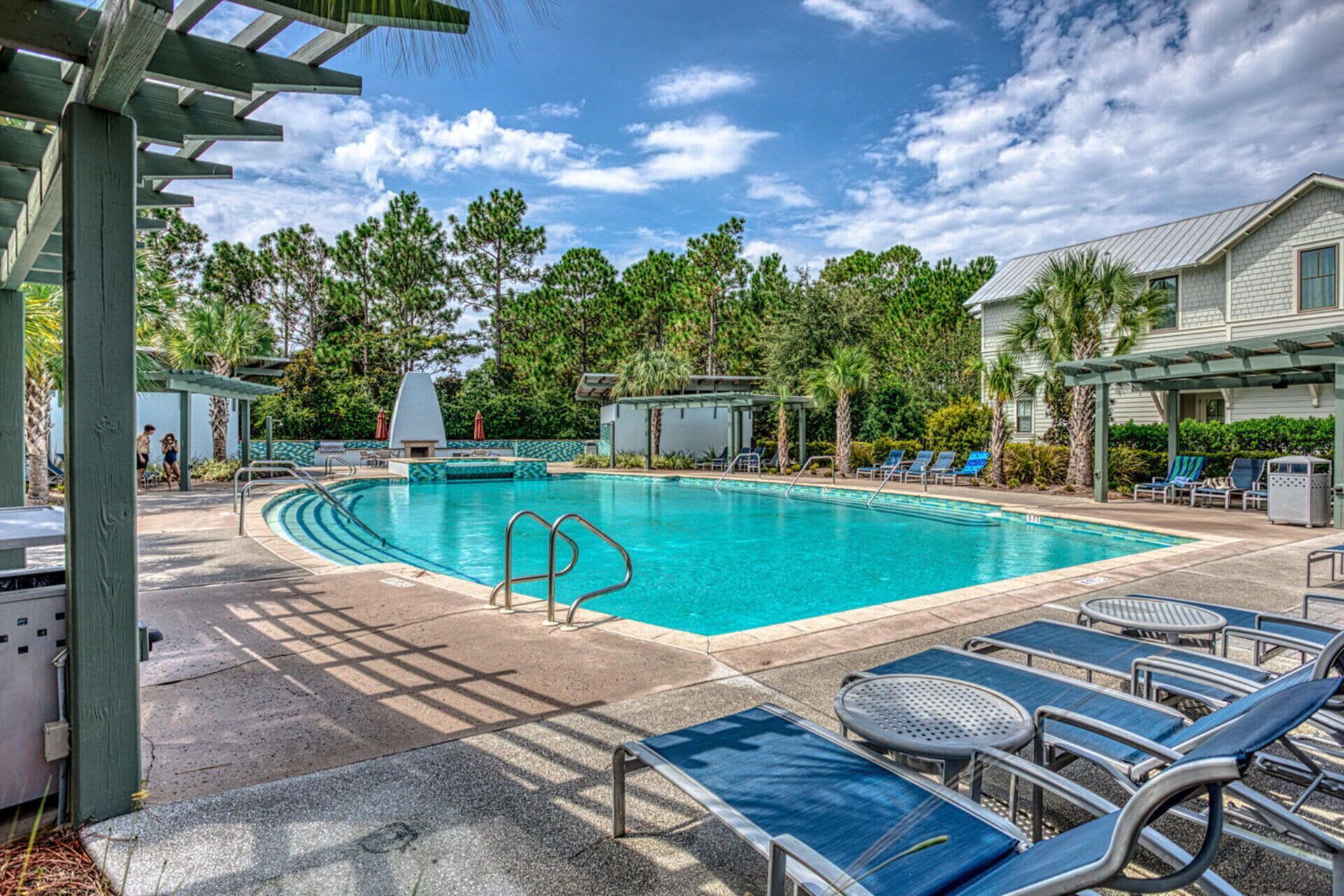 A swimming pool with lounge chairs and a covered area, surrounded by trees and a building.