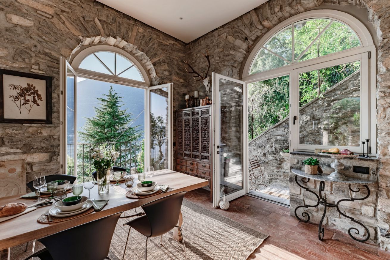 Un comedor con pared de piedra, una mesa de madera dispuesta para dos comensales y vistas al exterior con vegetación.