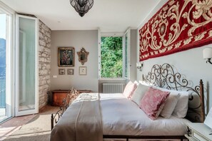 A bedroom with a bed, a window, a stone wall, and a view of greenery. - Villa La Solitaria - My Home in Como (Blevio)
