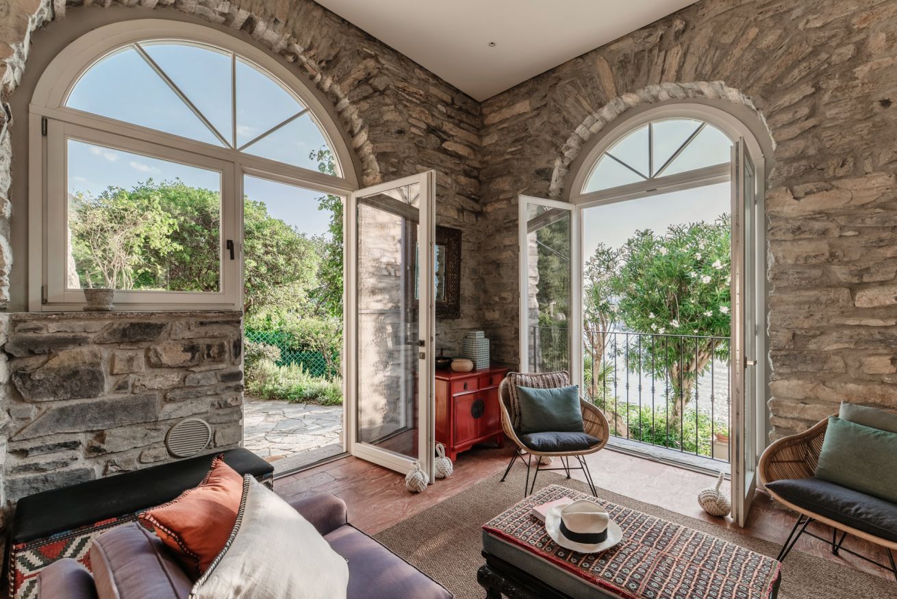 A room with stone walls, large arched windows, a sofa, a chair, and a table with a hat.