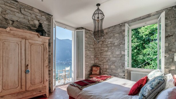 Ein Schlafzimmer mit Steinwand, einem Holzschrank, einem Bett mit Kissen, einem roten Teppich und Blick ins Grüne durch ein Fenster.
