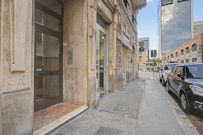 Une vue de rue avec un immeuble doté d’une grande porte vitrée, d’une grille métallique sur le trottoir et de voitures garées.