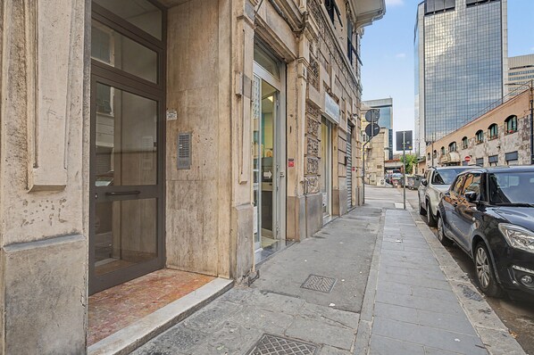 Una vista di strada con un edificio dotato di un'ampia porta in vetro, una grata metallica sul marciapiede e auto parcheggiate.