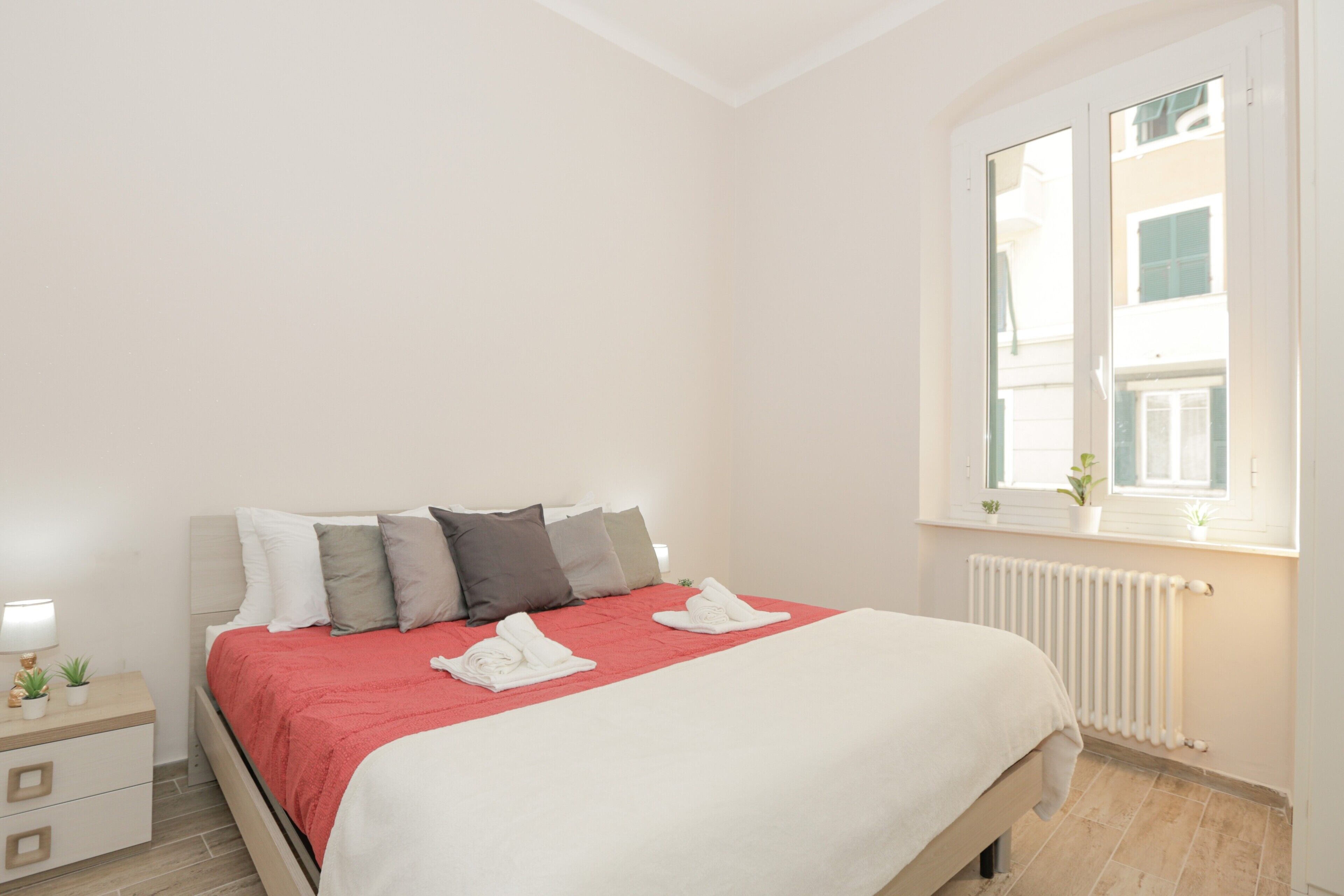 Une chambre à coucher avec un lit, des oreillers, une table de chevet, une lampe et une fenêtre donnant sur des immeubles.