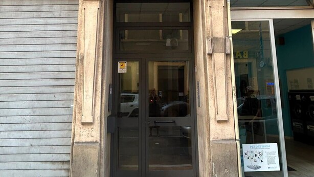Une entrée d’immeuble avec un volet fermé, une porte dotée d’un panneau de verre et une enseigne indiquant un service de nettoyage à sec pour animaux de compagnie.