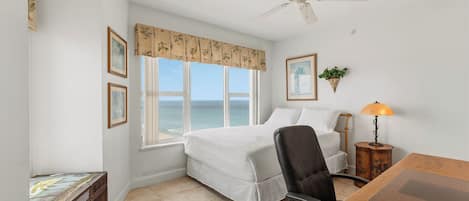 Un dormitorio con cama, una silla, un escritorio, una lámpara, un ventilador de techo y una ventana con vista al mar.